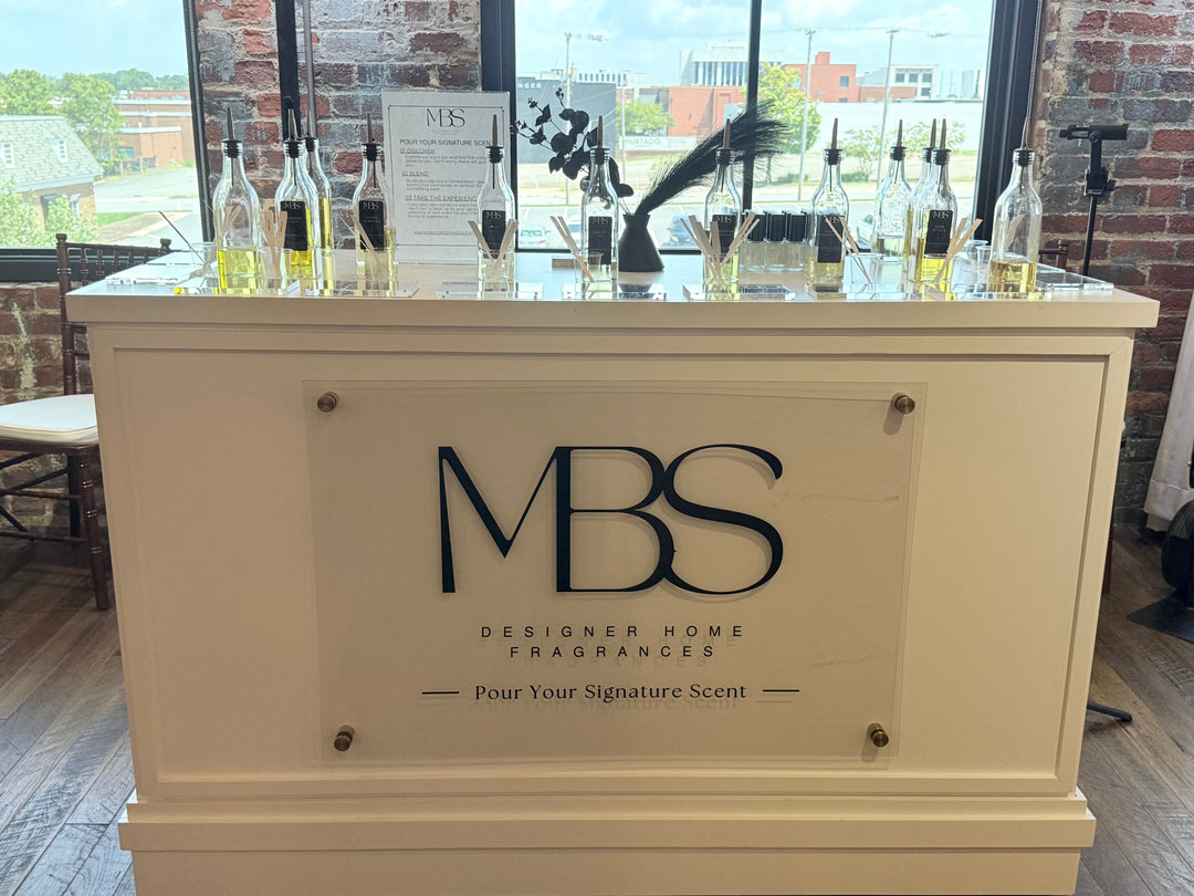 A white MBS Designer Home Fragrances counter display stand in a bright indoor space with brick walls and large windows. Multiple fragrance bottles are arranged on top, and the front panel features the MBS logo and the tagline 'Pour Your Signature Scent'. Candle making. Raleigh Candles. Raleigh Candle experiences.